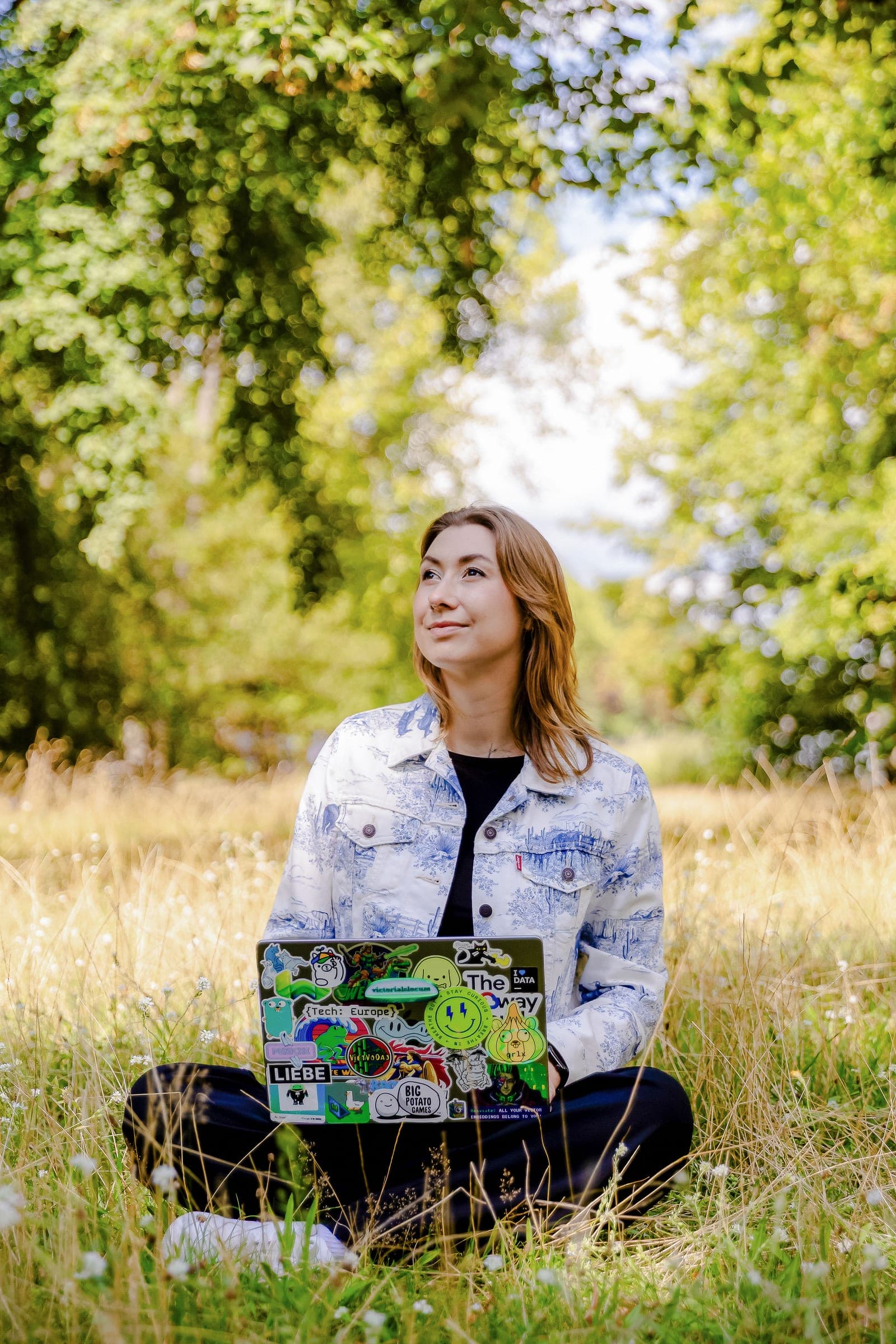 Victoria sitting cross-legged in a sunlit meadow, a sticker-covered laptop open on her knees, looking up into the trees.