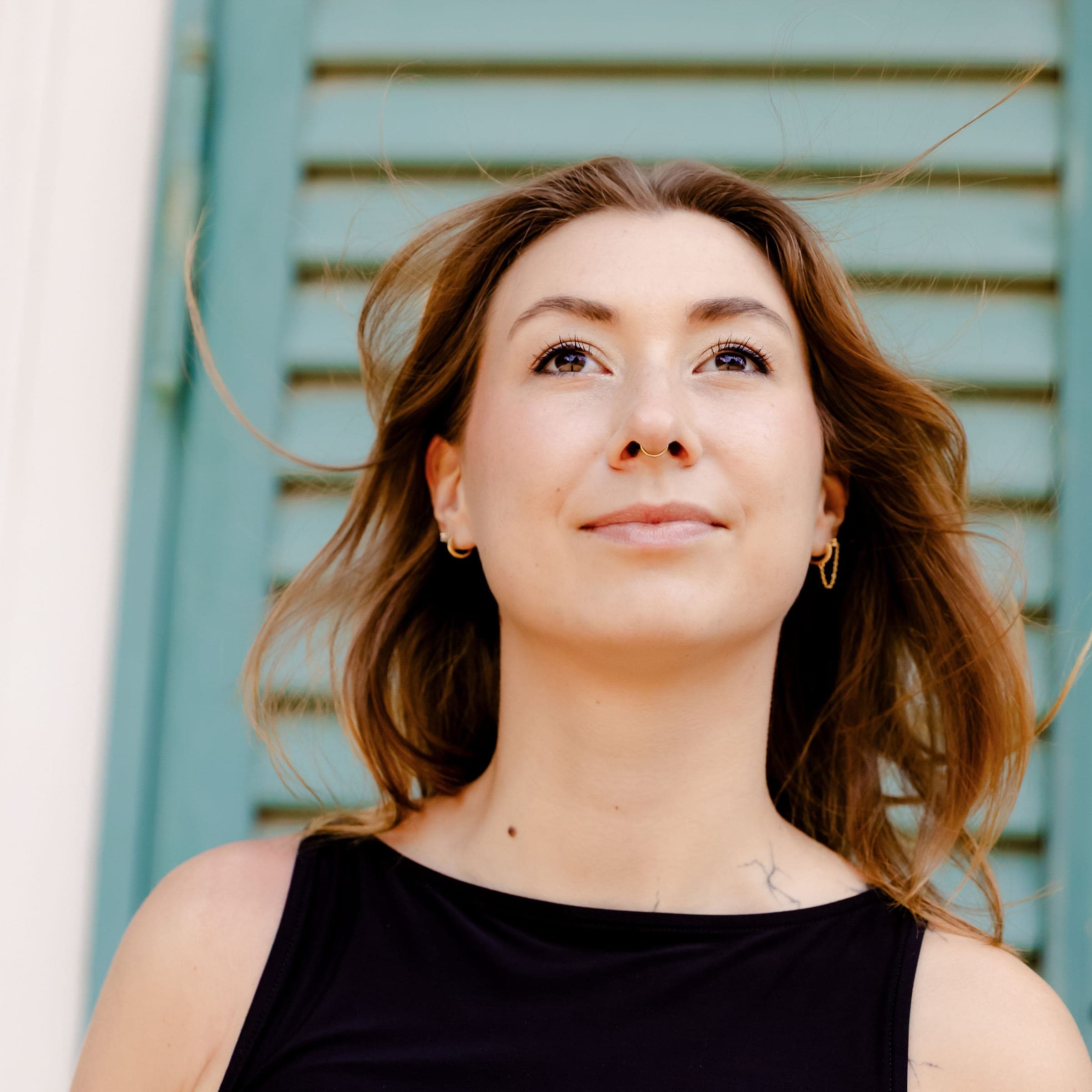 Victoria in a black top, chin raised, looking off to one side, against a pale green shuttered wall.
