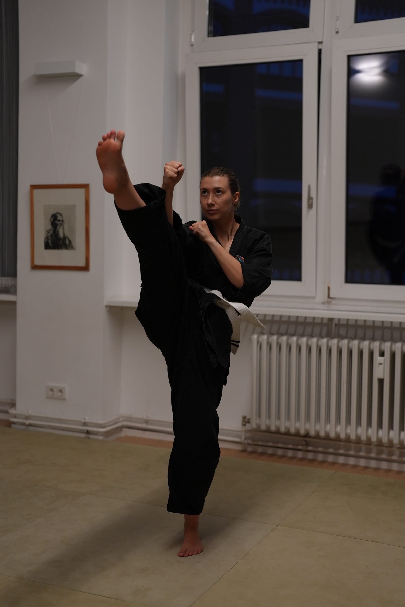 Victoria mid-kick in a black martial arts uniform, leg extended high, in a room with tall windows.