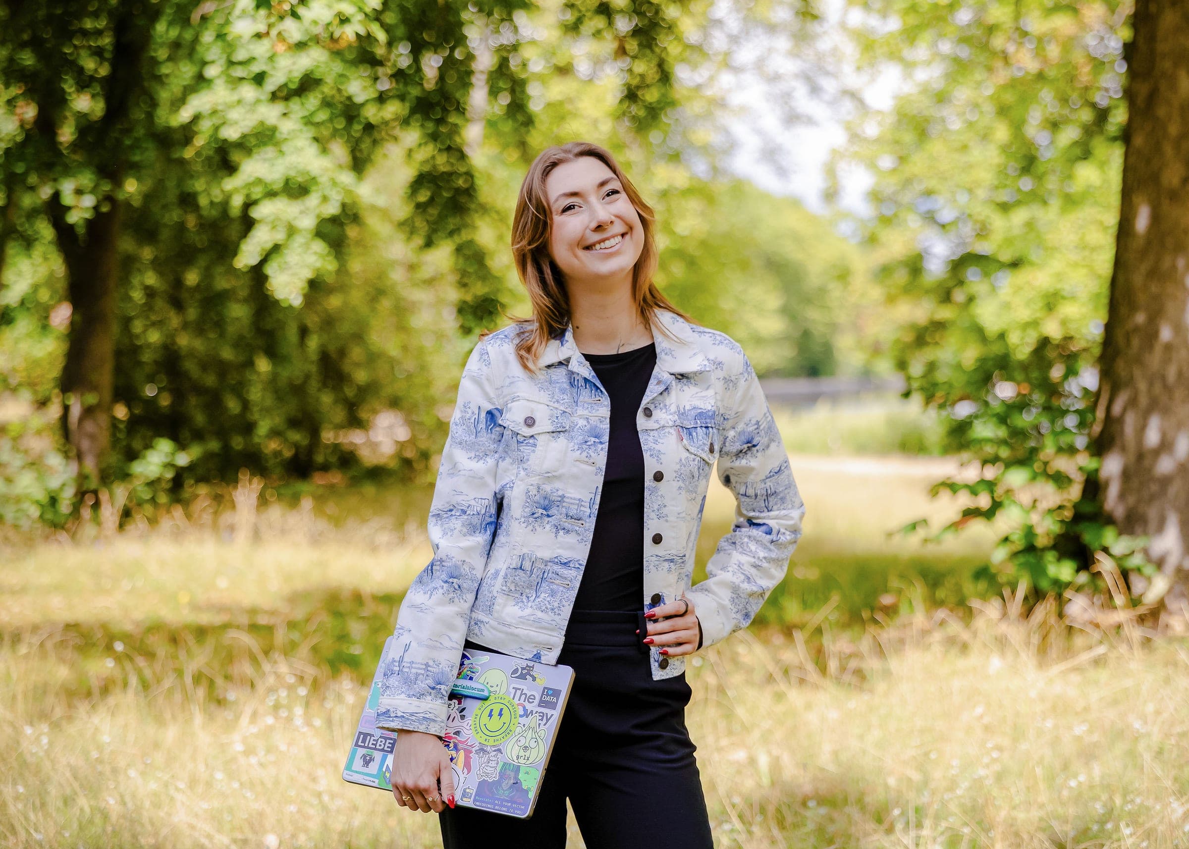 Victoria standing in tall meadow grass holding a sticker-covered laptop, laughing.