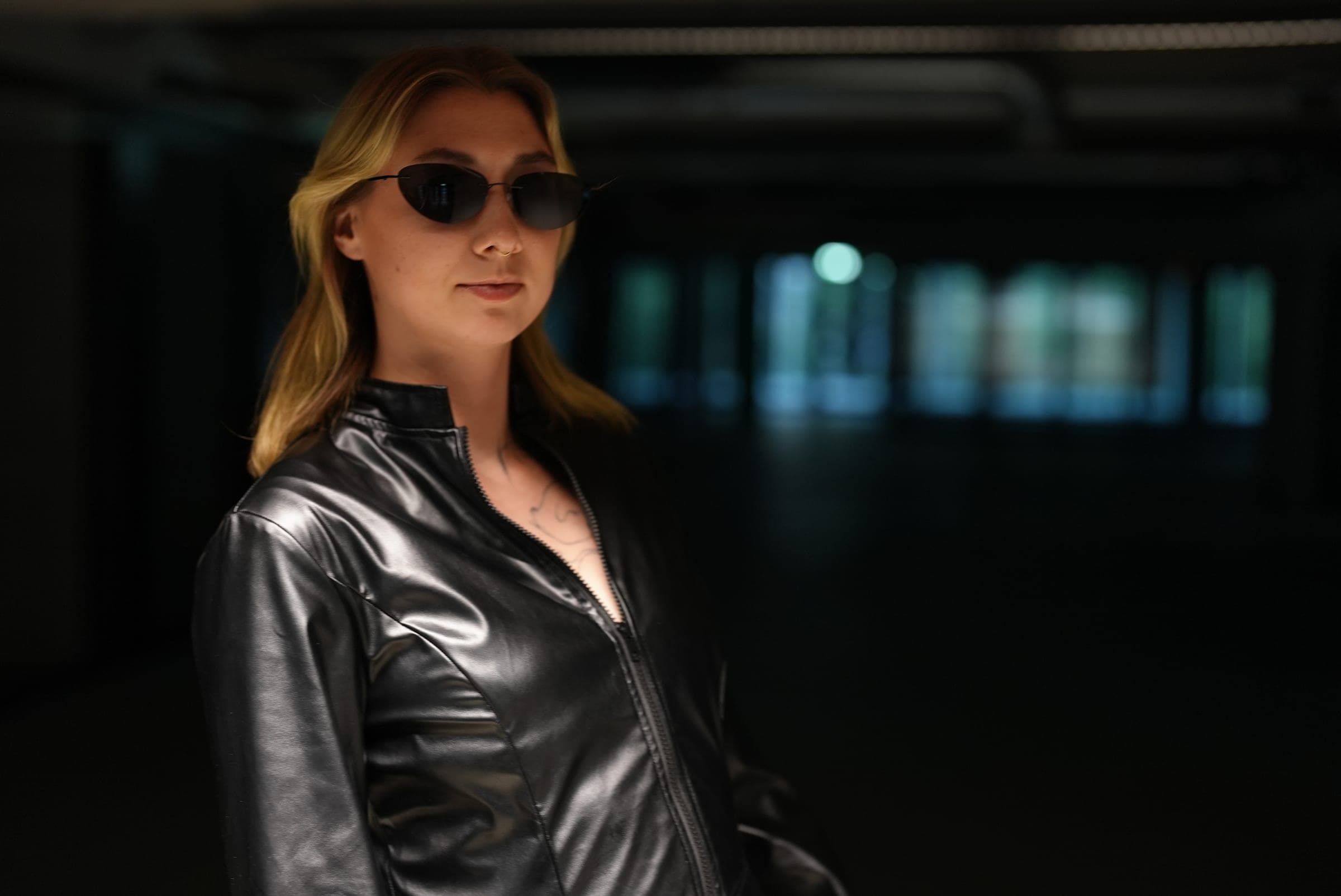Victoria in a black leather jacket and dark sunglasses in a dim industrial space.